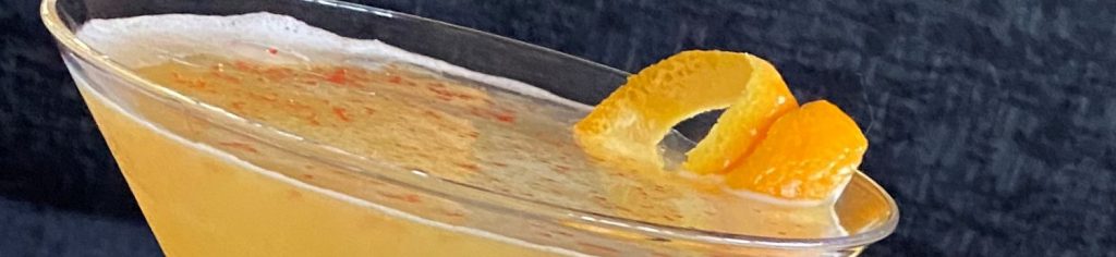 Chili Citrus Sour: Gold cocktail in a martini glass with an orange peel curl and sprinkling of red chili on the surface