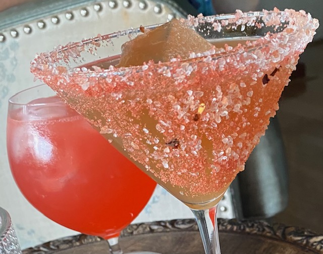 Hot Cacao Margarita: Pale pink cocktail in a martini glass completely crusted in pink cranberry salt; a pink cocktail in the background in a goblet