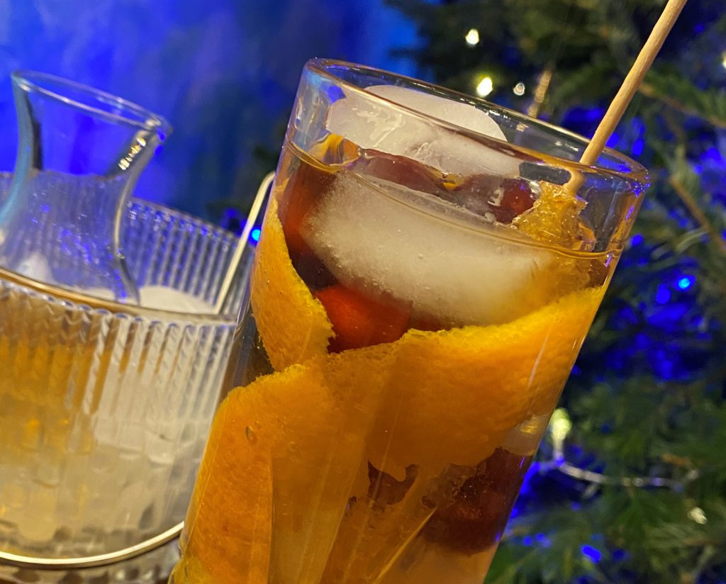 Orange Ginger Old Fashioned: Clear beverage in a highball glass with orange peel ribbons, cranberries, and a gold sugar stick