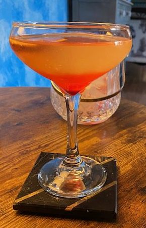 Ruby Diamond Elixir: White cocktail with oink hue in a martini glass with a thin tangerine slice