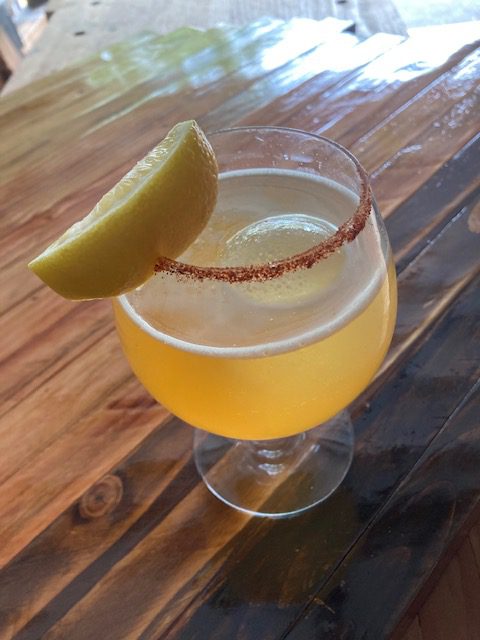 Workshop Beer Cocktail: Golden drink in a goblet with large ice sphere and lemon wedge, against a workshop background