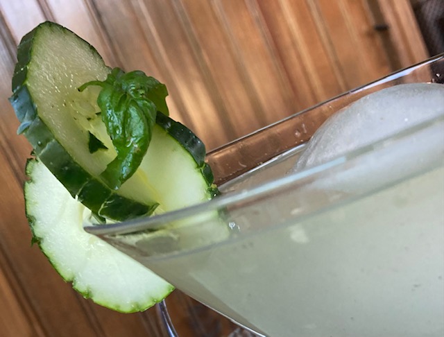 Cucumber Experience: Pale green cocktail with ice sphere in a martini glass garnished with scored cucumber slices and basil leaf