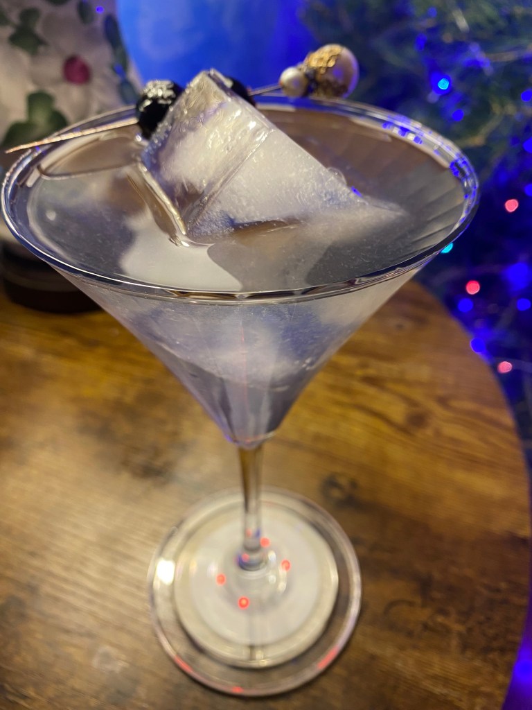 Clear cocktail in a martini glass with large ice cube and speared Luxardo cherries