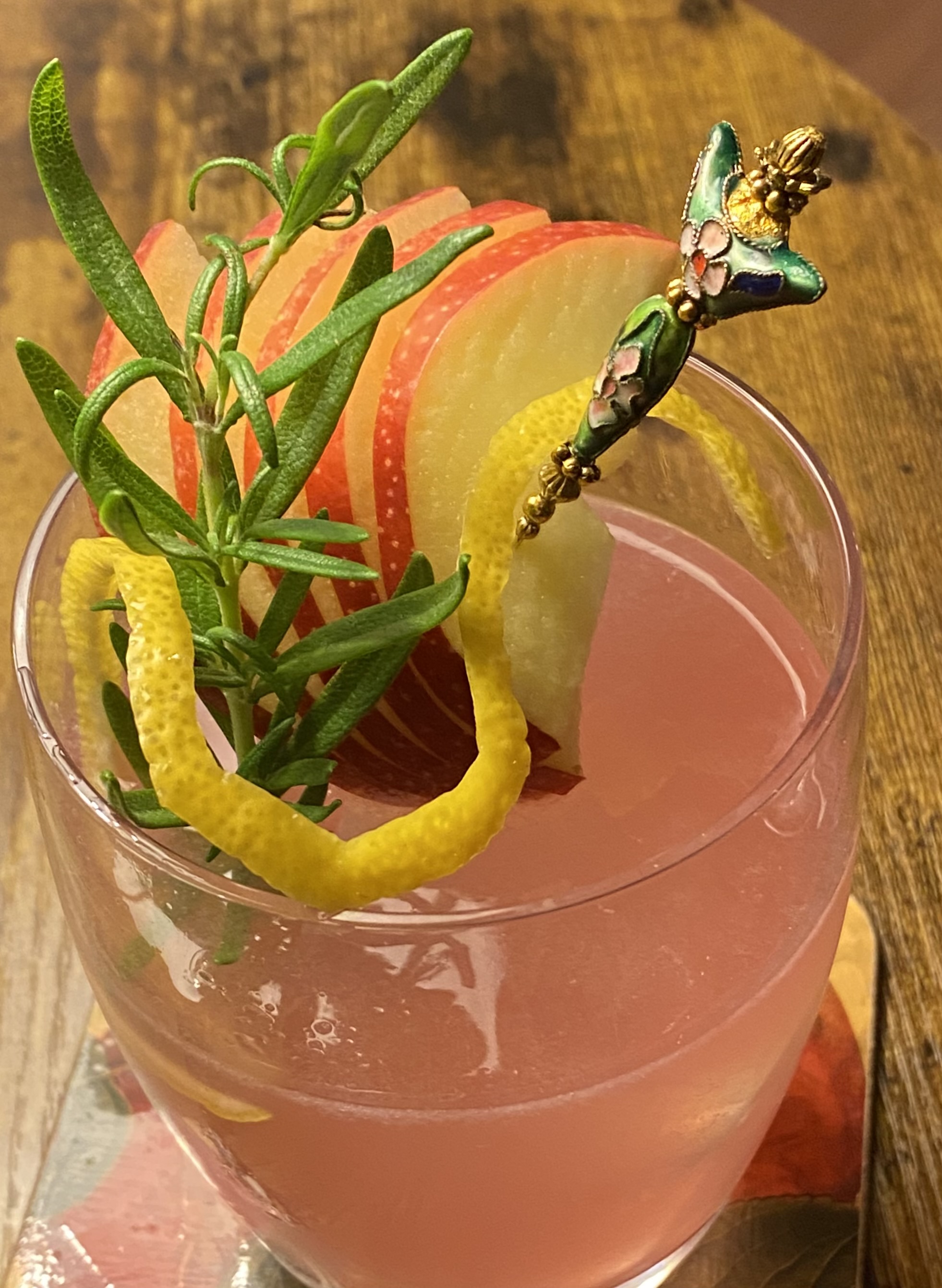 Pale pink drink in old-fashioned glass, apple-rosemary-lemon curl garnish on a hatpin, no ice