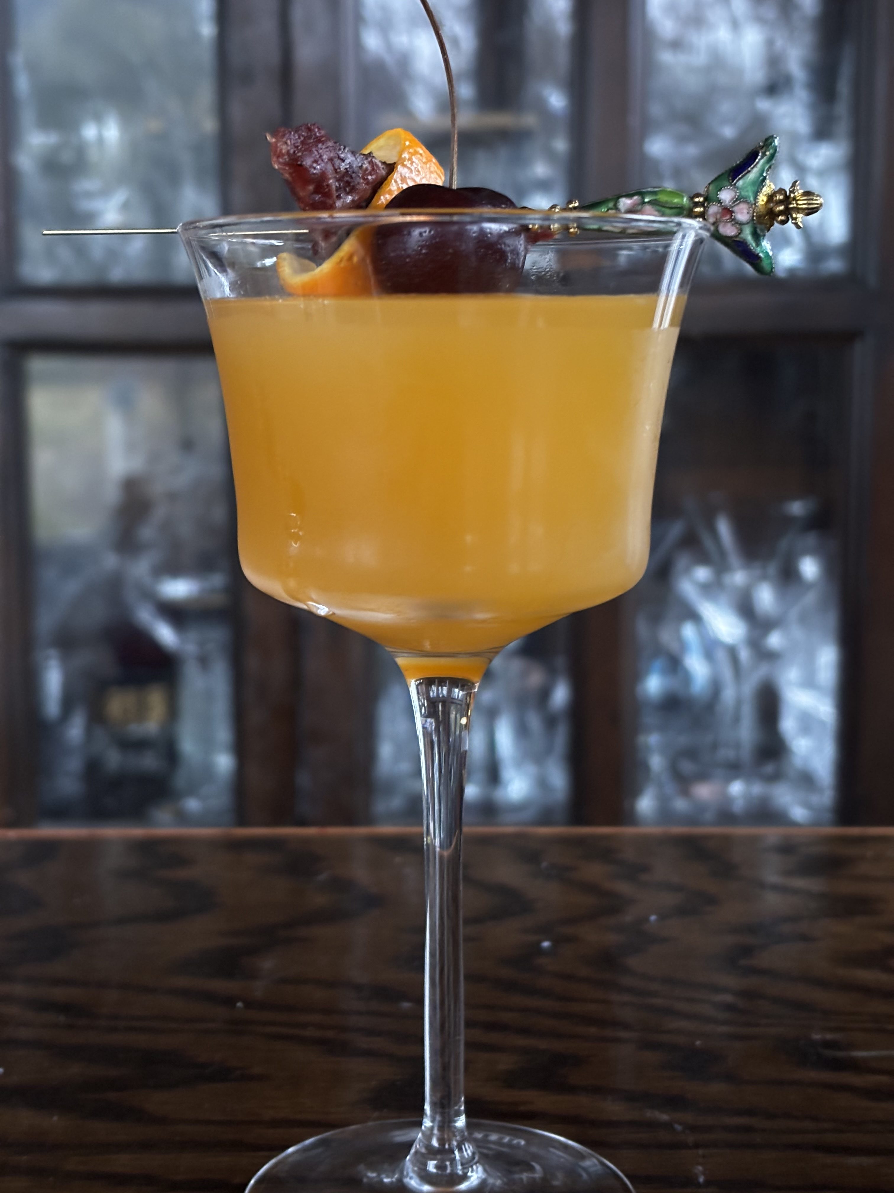Golden cocktail in Nick and Nora glass garnished with a fresh cherry, orange peel, and beef jerky.