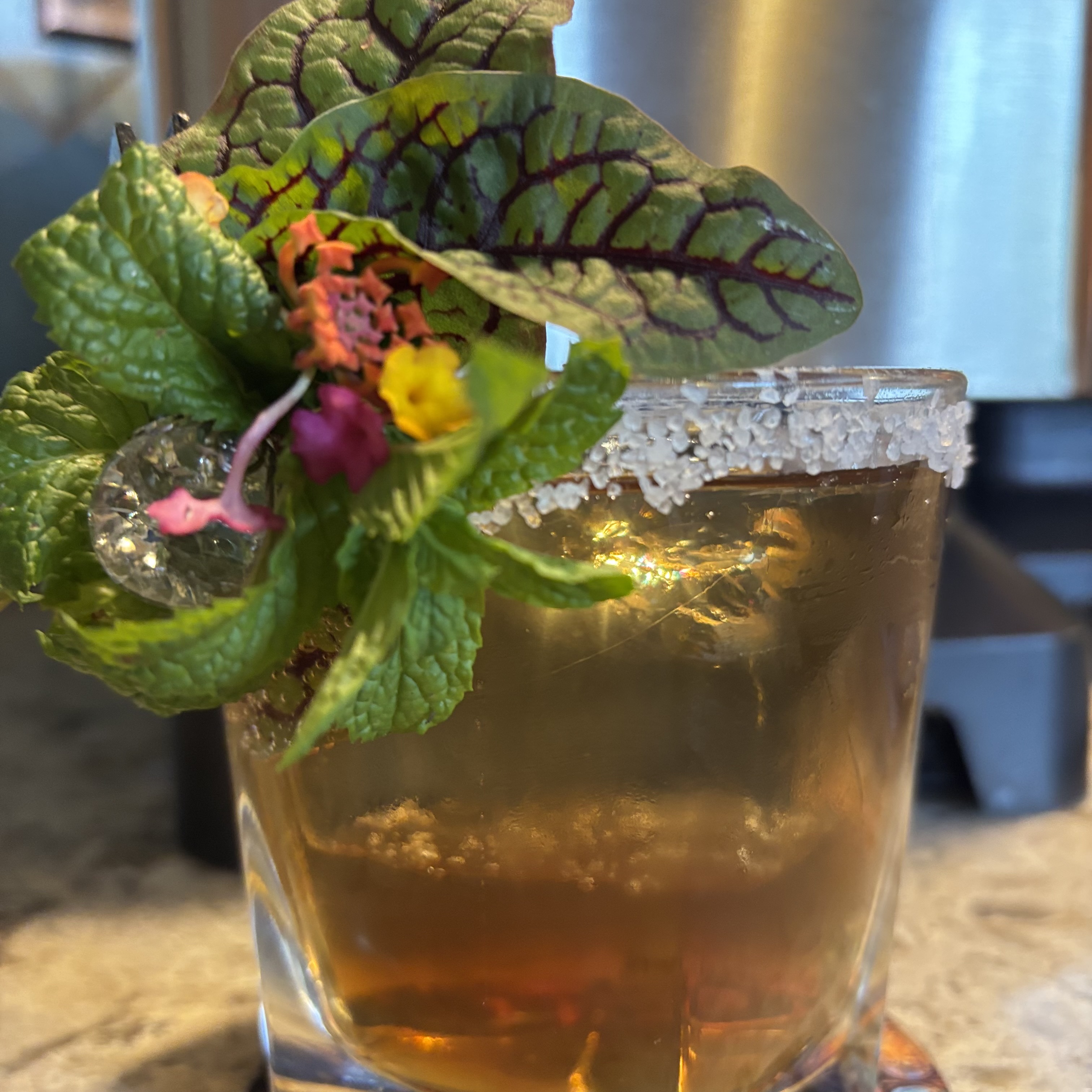 Bantanga: Light brown cocktail in an old-fashioned glass with a large cube, rimmed with salt, garnished with flowers and herbs
