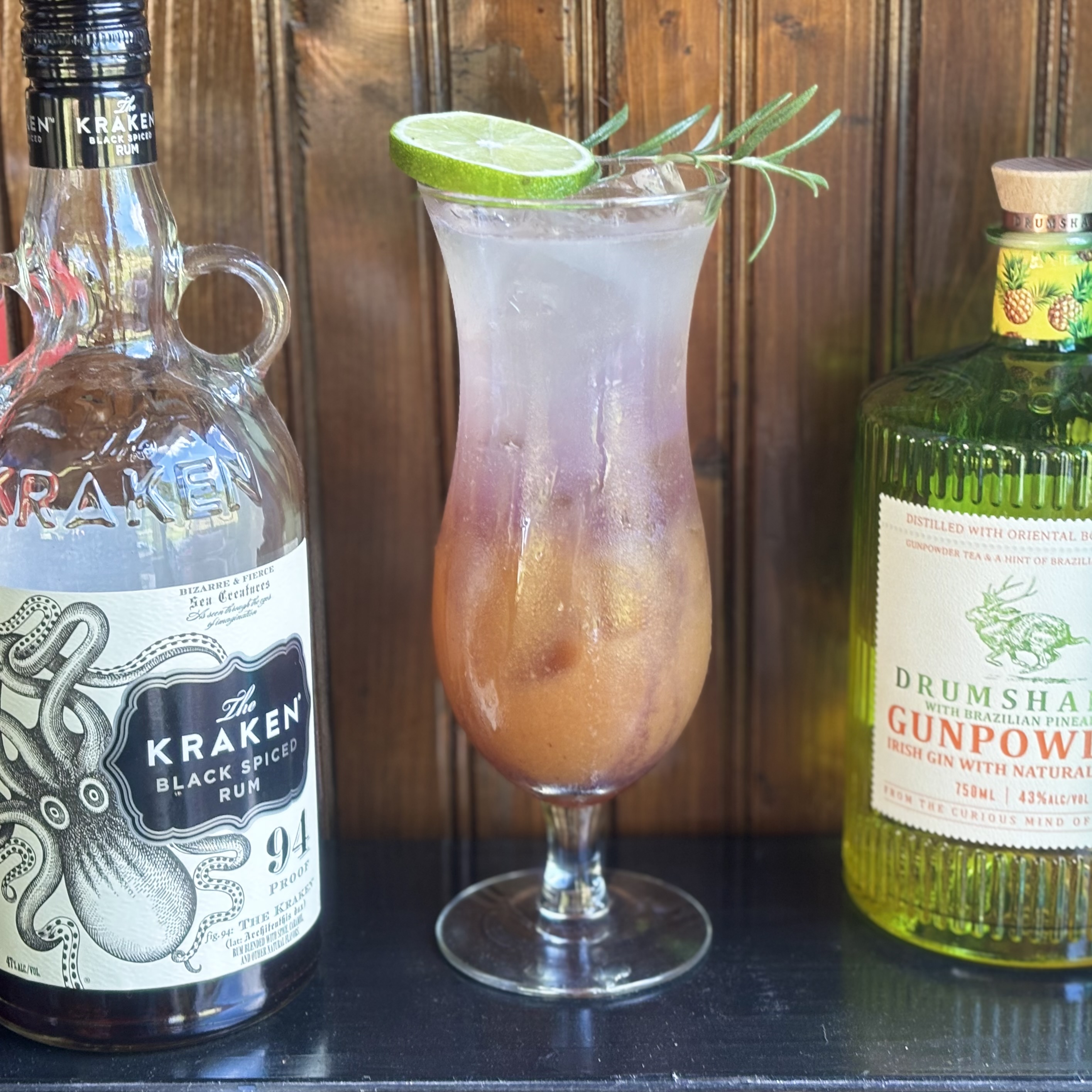 Dichotomy: Multi-layer drink with orange, purple, and white sections in a hurricane glass garnished with a lime wheel and rosemary sprig
